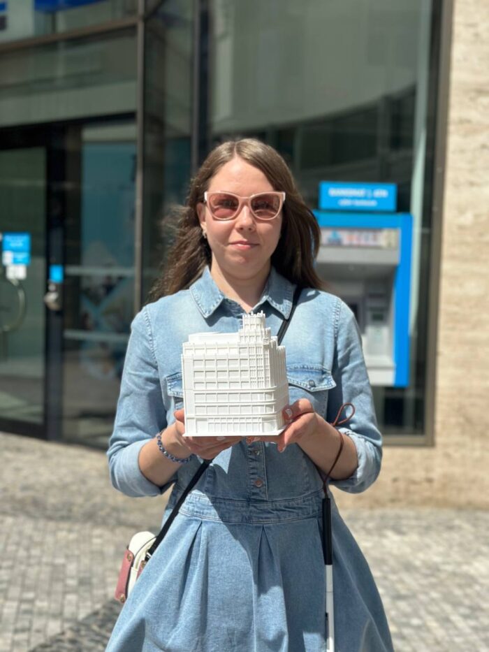 Na fotografii je mladá žena s dlouhými hnědými vlasy, oblečená v světle modrých džínových šatech a růžových slunečních brýlích. Stojí před moderní budovou s velkými skleněnými okny a v rukou drží bílý model budovy. V pozadí je vidět bankomat a část nápisu „BANKOMAT 24/7“. Žena má také bílou hůl, což naznačuje, že je nevidomá nebo slabozraká.